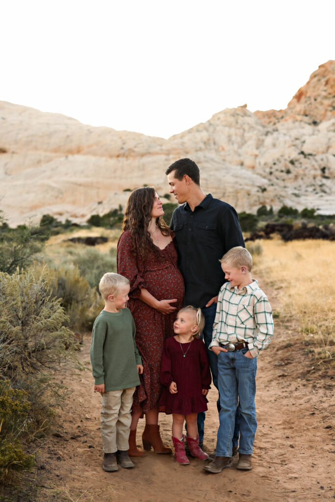 A family of 5 looks at each other while mom cradles her baby belly during family photos in Snow Canyon State Park. The white rocks in the background make a beautiful backdrop.