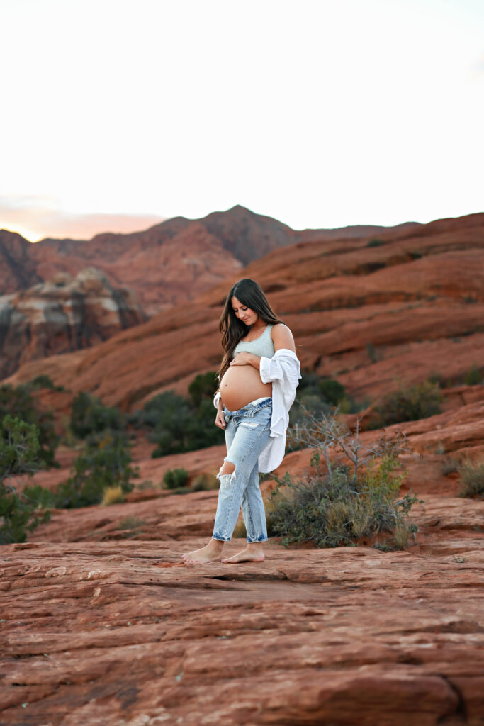 Expectant mother standing on red rock formations at Snow Canyon State Park in Southern Utah, surrounded by desert landscape and dramatic cliffs.