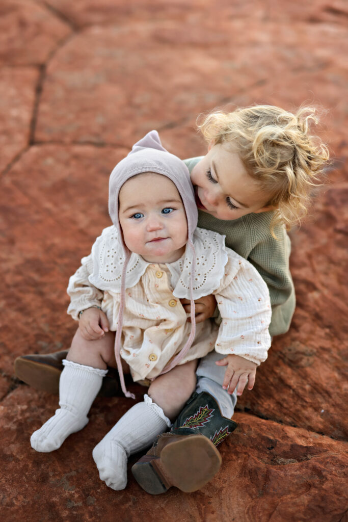 Cute baby and toddler sitting together on the red rocks along the Butterfly Trail in Snow Canyon State Park, surrounded by vibrant desert landscape and sandstone formations.