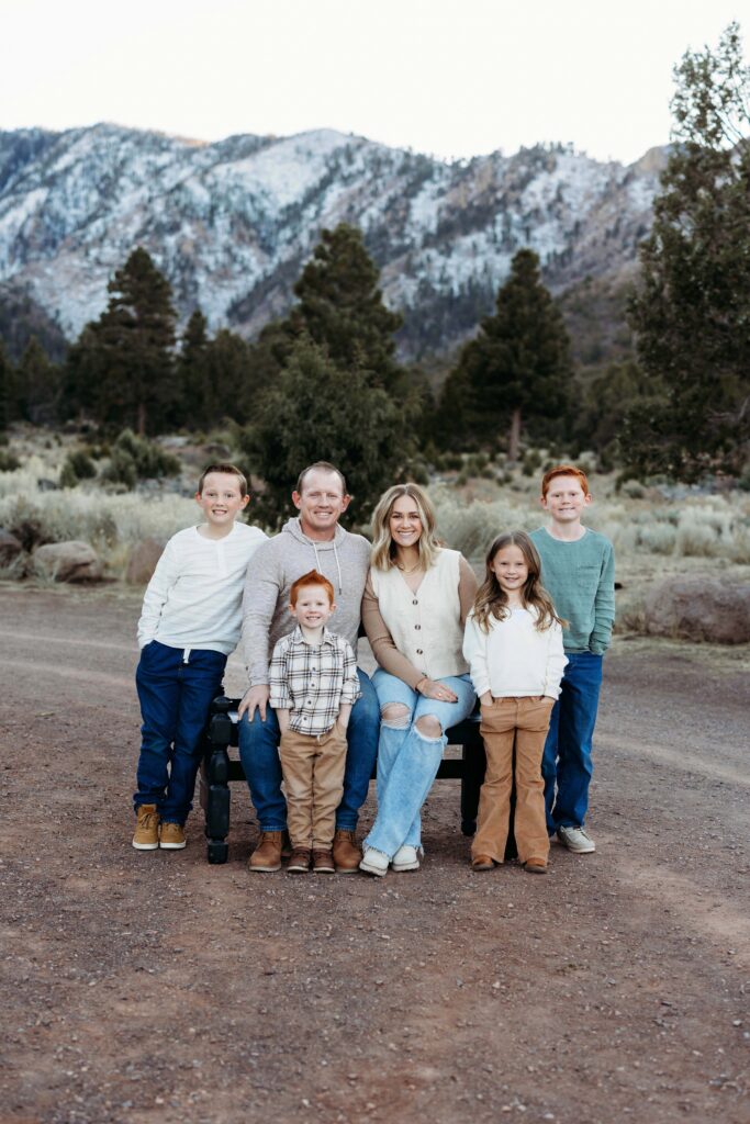 Family of six smiling and posing for a family portrait in Pine Valley, Utah with snowy mountains and pine trees behind them.