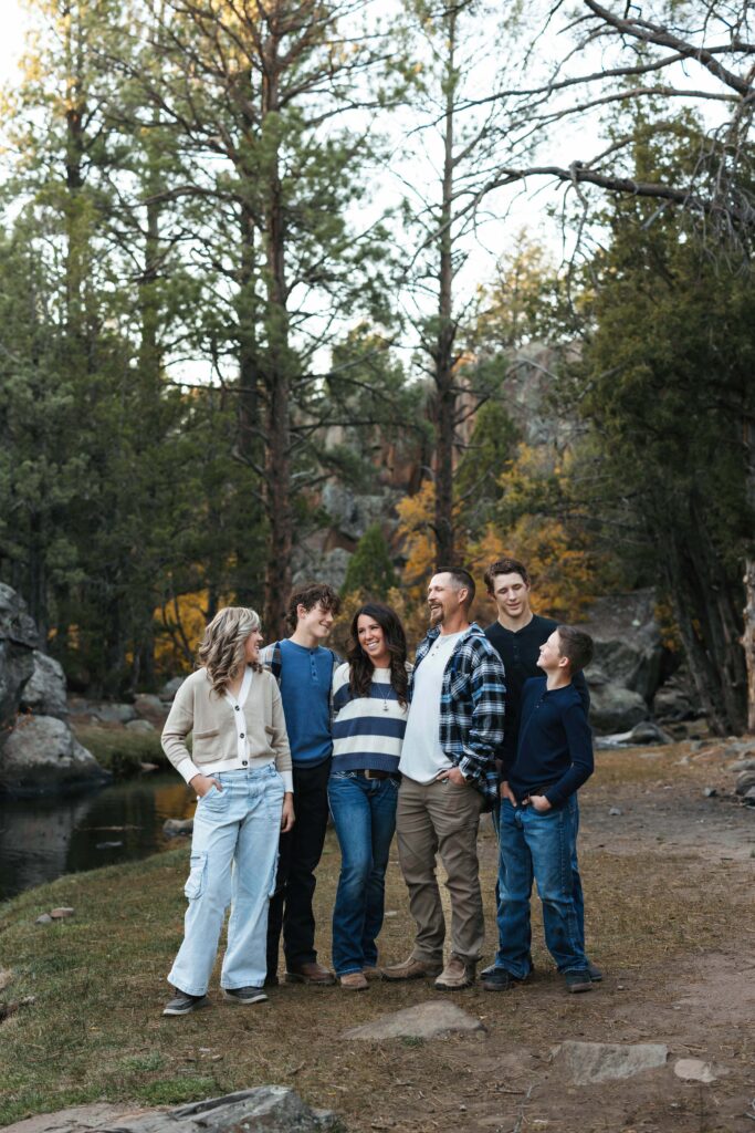 Family of six standing and laughing together next to a river and among tall pine trees in Pine Vally, Utah dressed in cozy fall outfits.