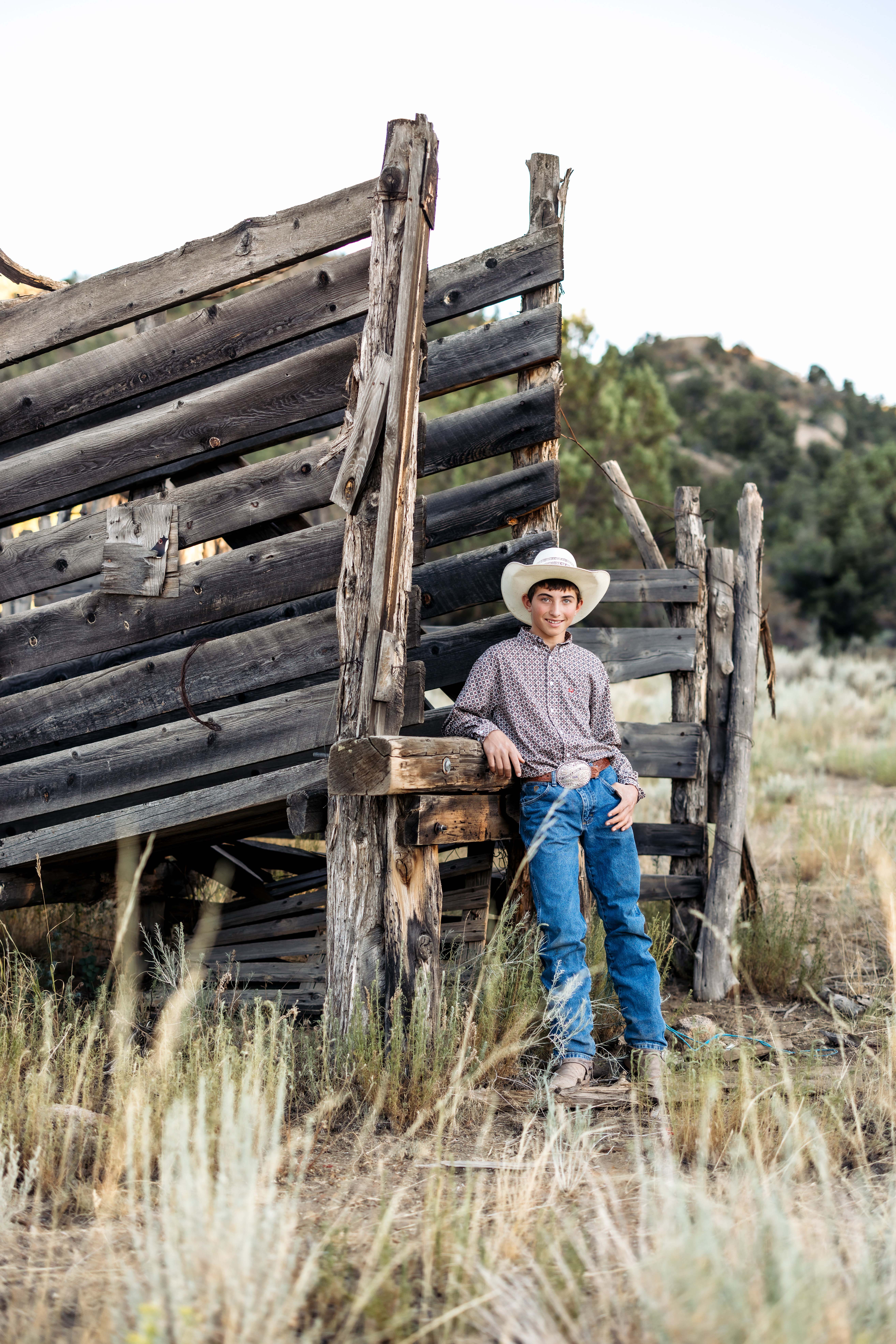 A cowboy poses against an old corral while wearing a cowboy hat and western wear