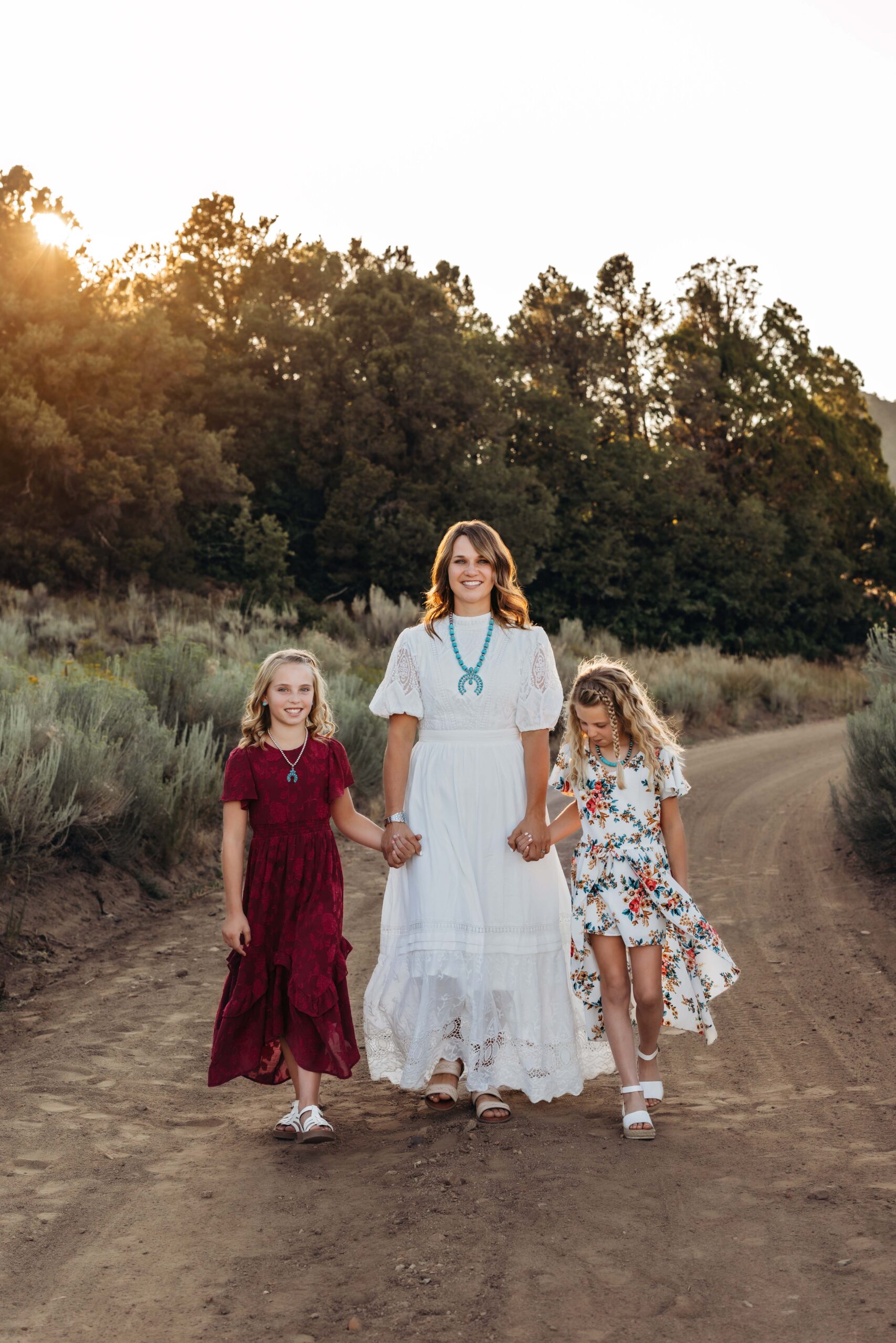 A mom holds hands with her two daughters as they walk down a dirt road in the golden evening light