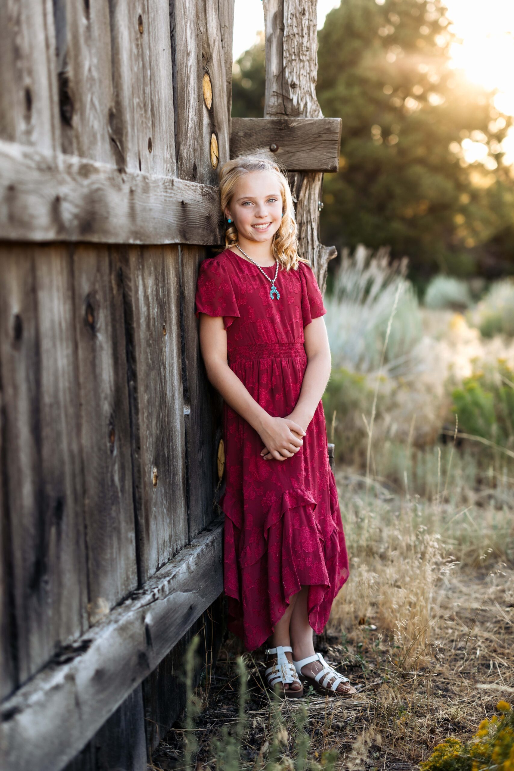 A pretty girl in a long red dress stands against an old wooden fence while the soft golden light glows behind her