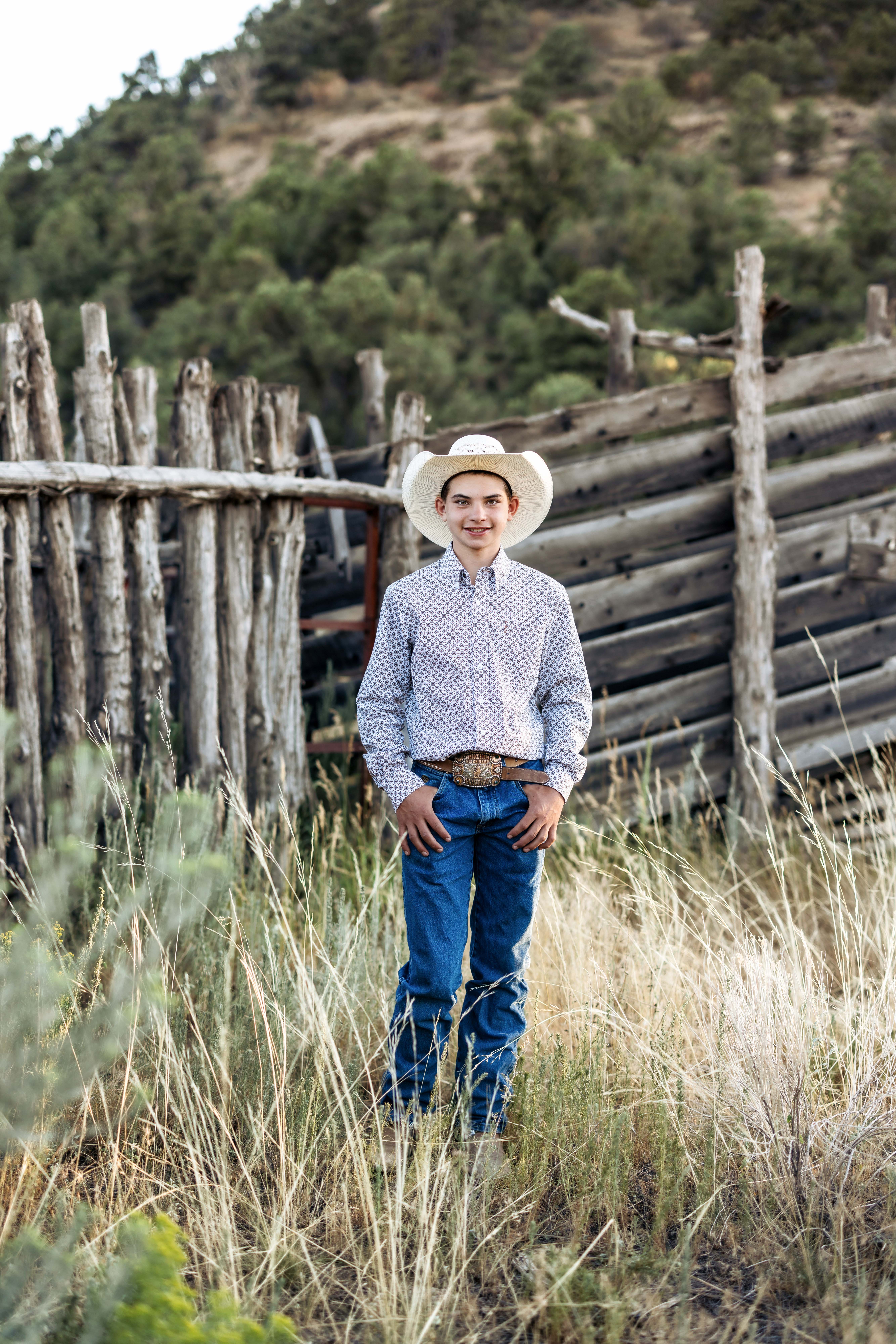 A boy stands and poses for a portrait in front of an old corral while dressed in western clothes