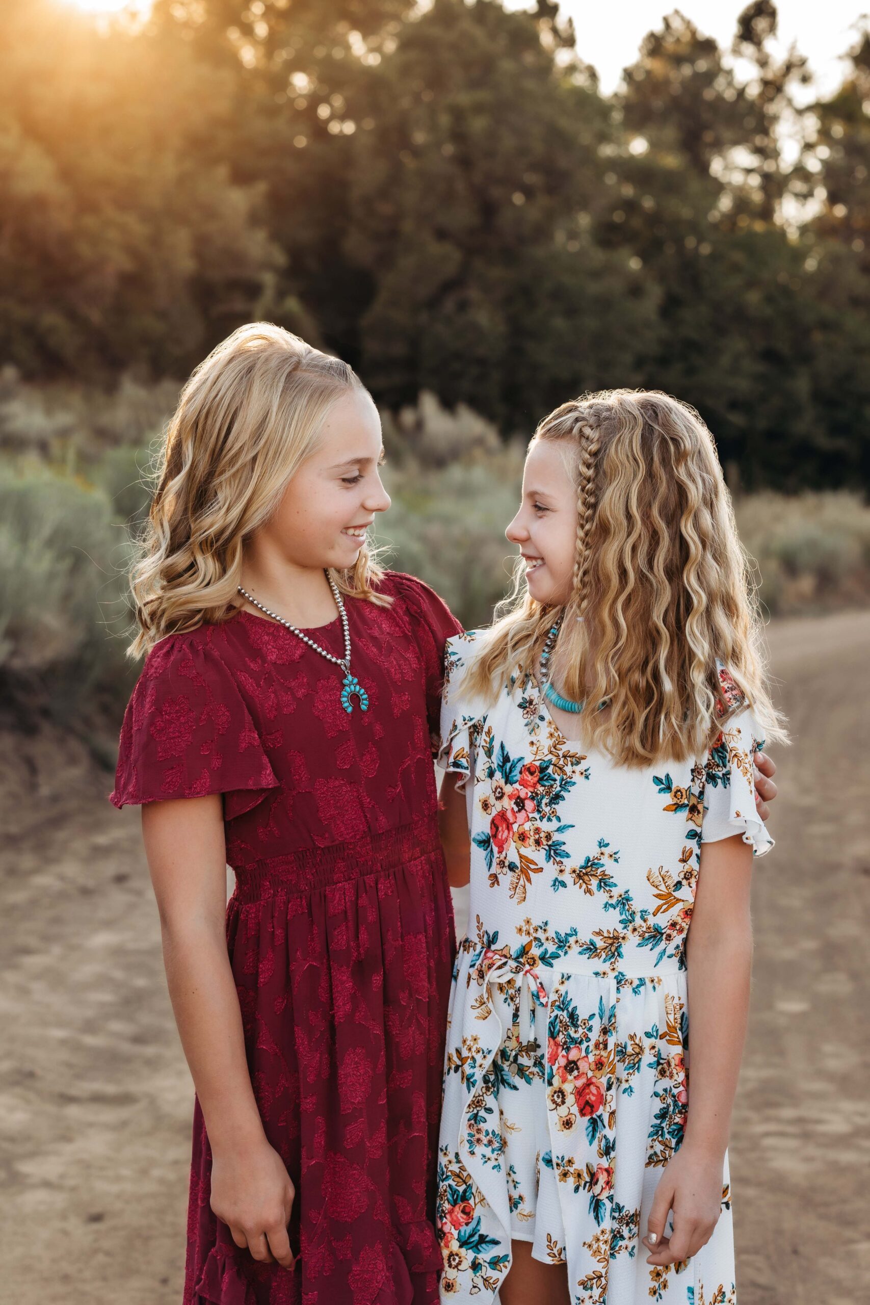 Two sister look at each other and smile with trees behind them and the soft glow of the golden sun