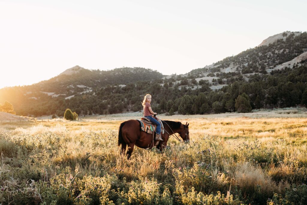 A girl sits on a horse in an open field with golden light and hills in the distance