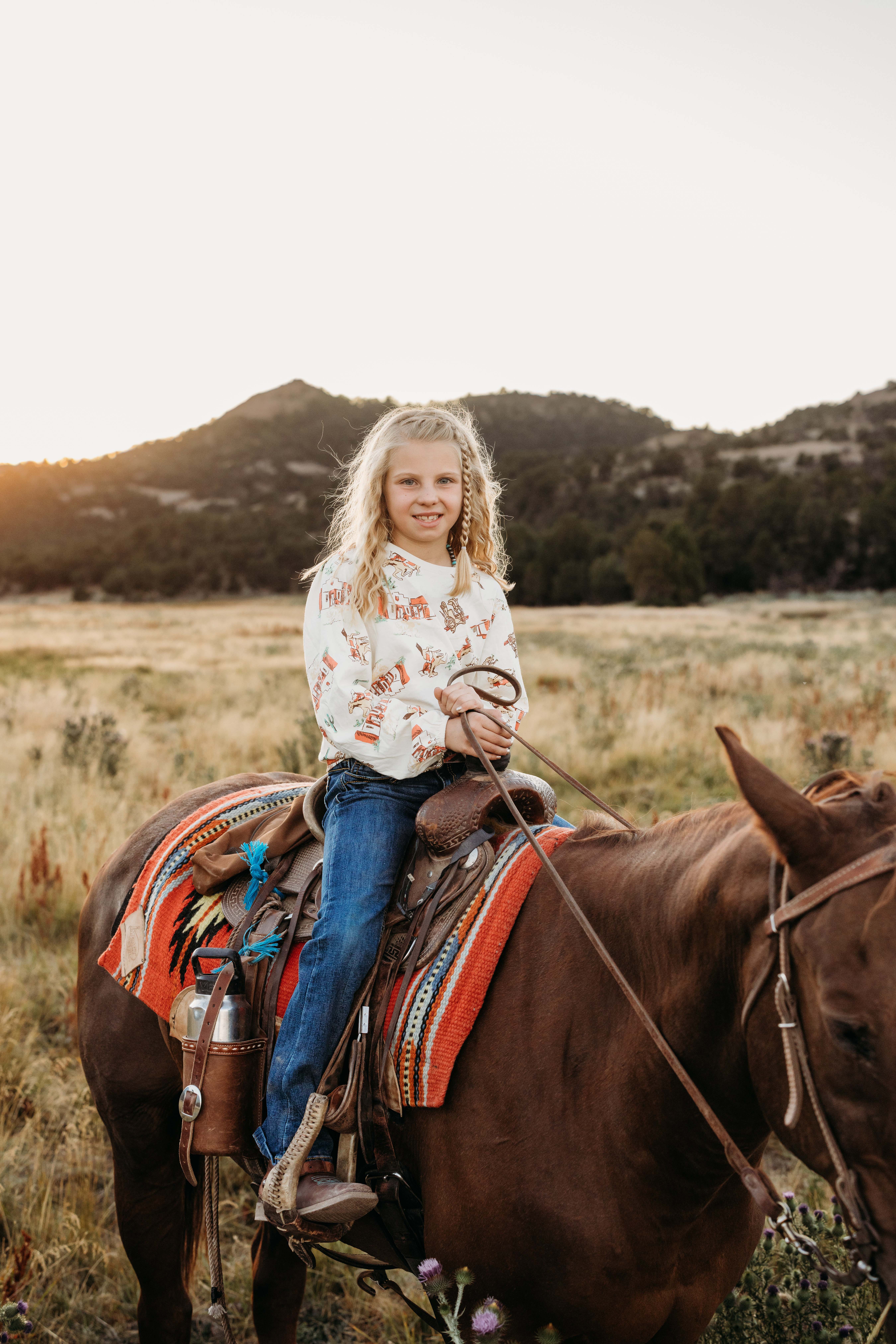 A girl poses for a picture while riding her horse with beautiful fields behind and soft golden light