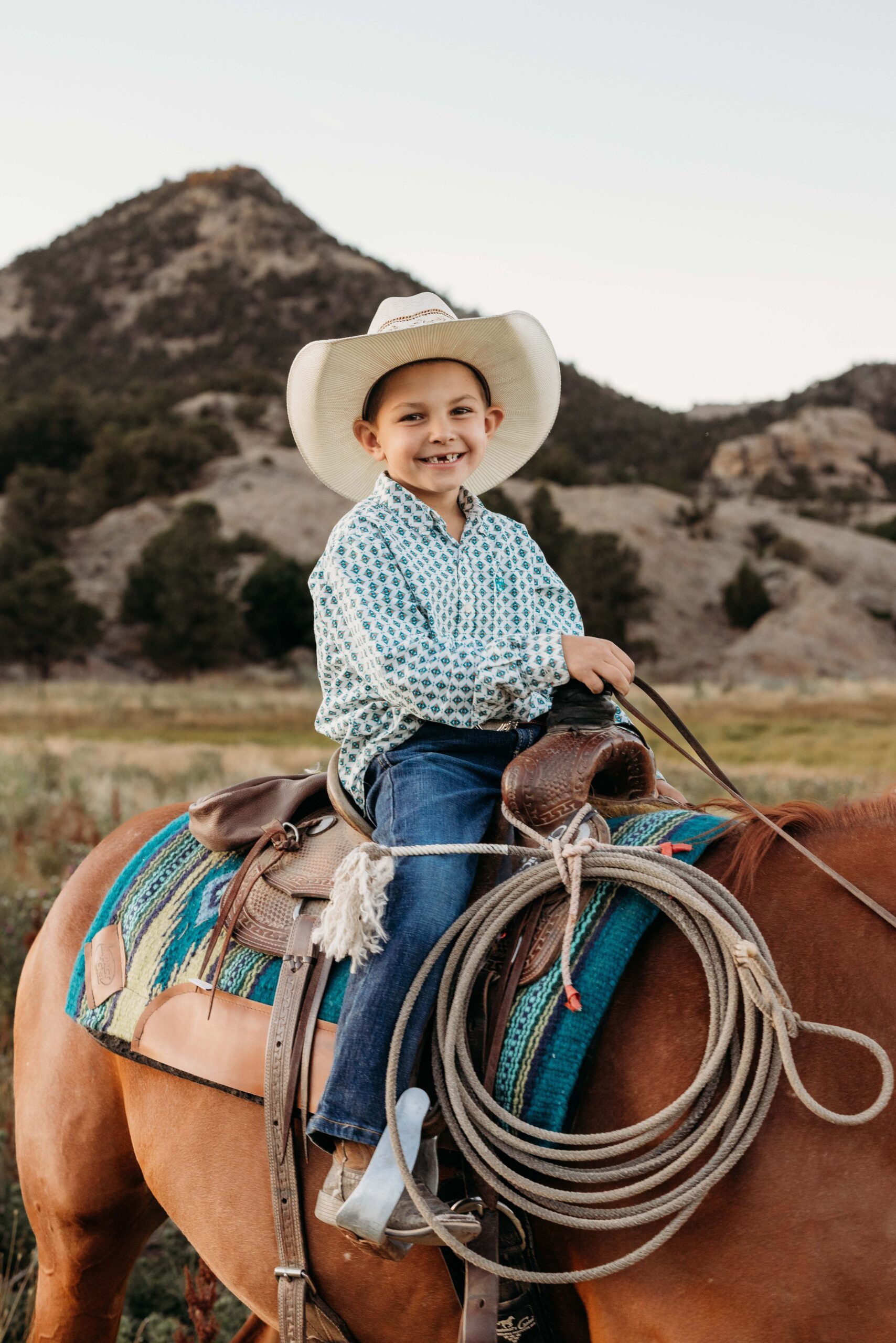 A little boy sits in his saddle on top his his horse with his lasso at his side and his cowboy hat on his head