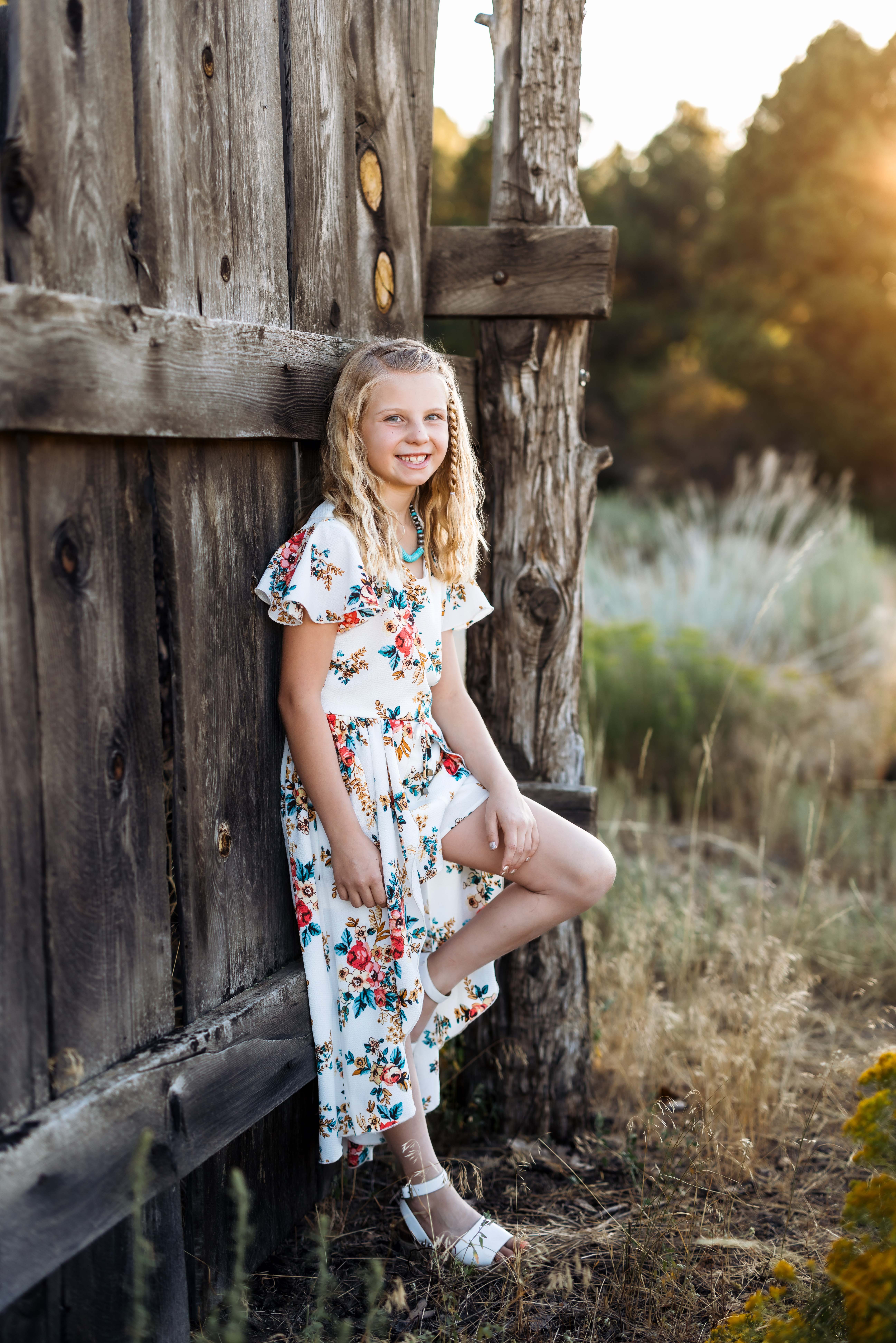 A girl wears a white floral dress and stands against an old weathered coral for a portrait