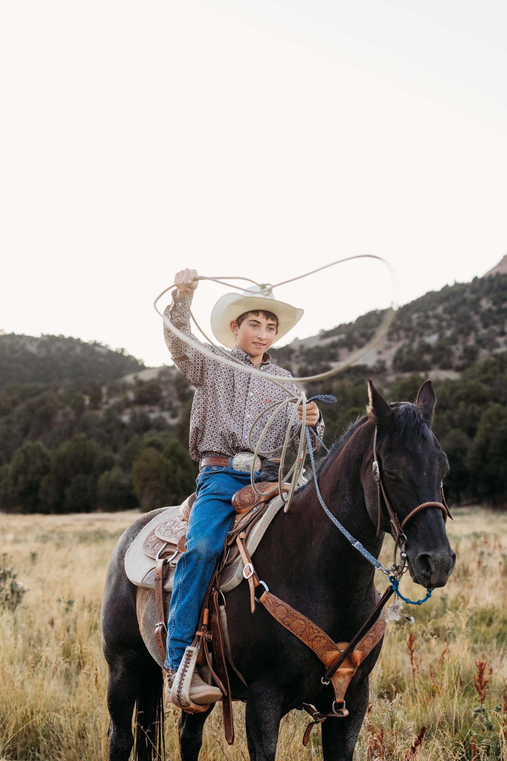 Cowboy swinging a lasso while sitting on top of his horse in an open field with mountain scenery behind him