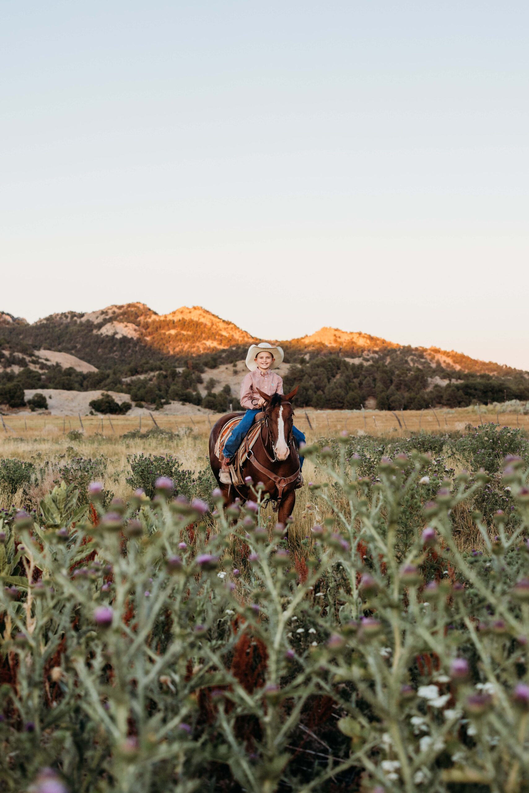 A boy smiles as he rides his horse through a field of cactus and yellow grass with the sun and the mountains behind him
