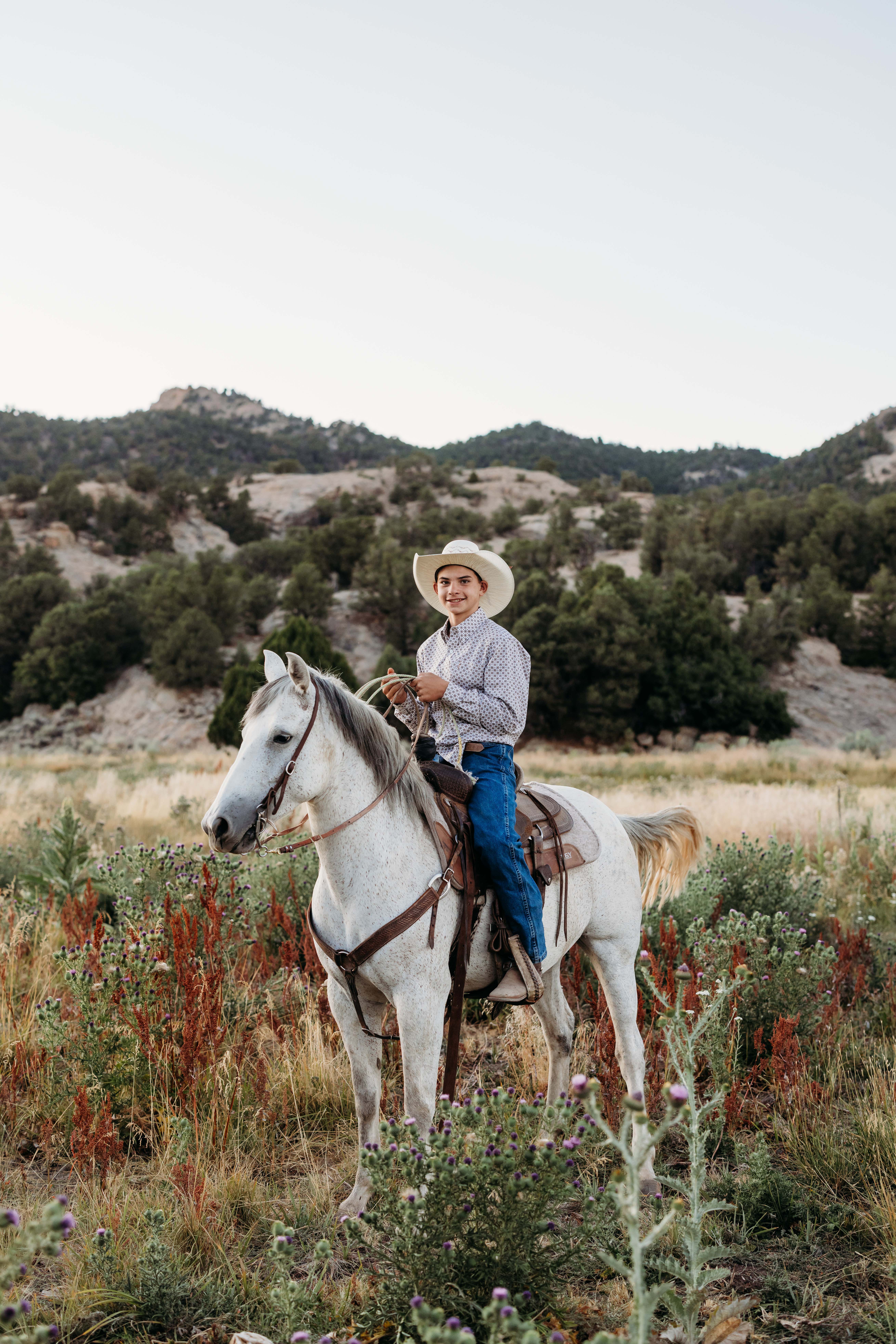 A teenage boy rides his beautiful white horse in an open field next to a jaw dropping mountain landscape