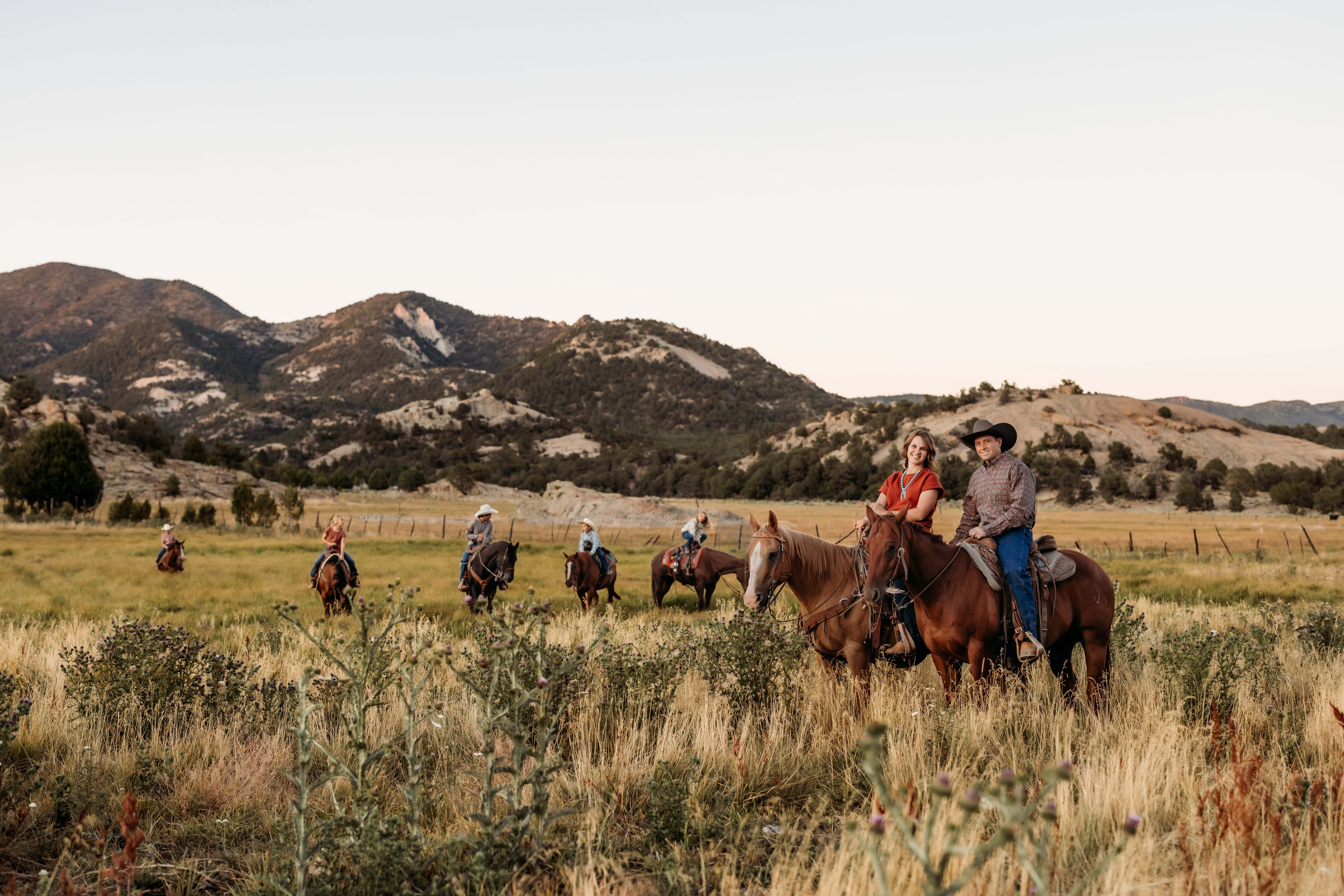 A couple sits on horseback and smiles for a photo as their kids ride horses in the background behind them