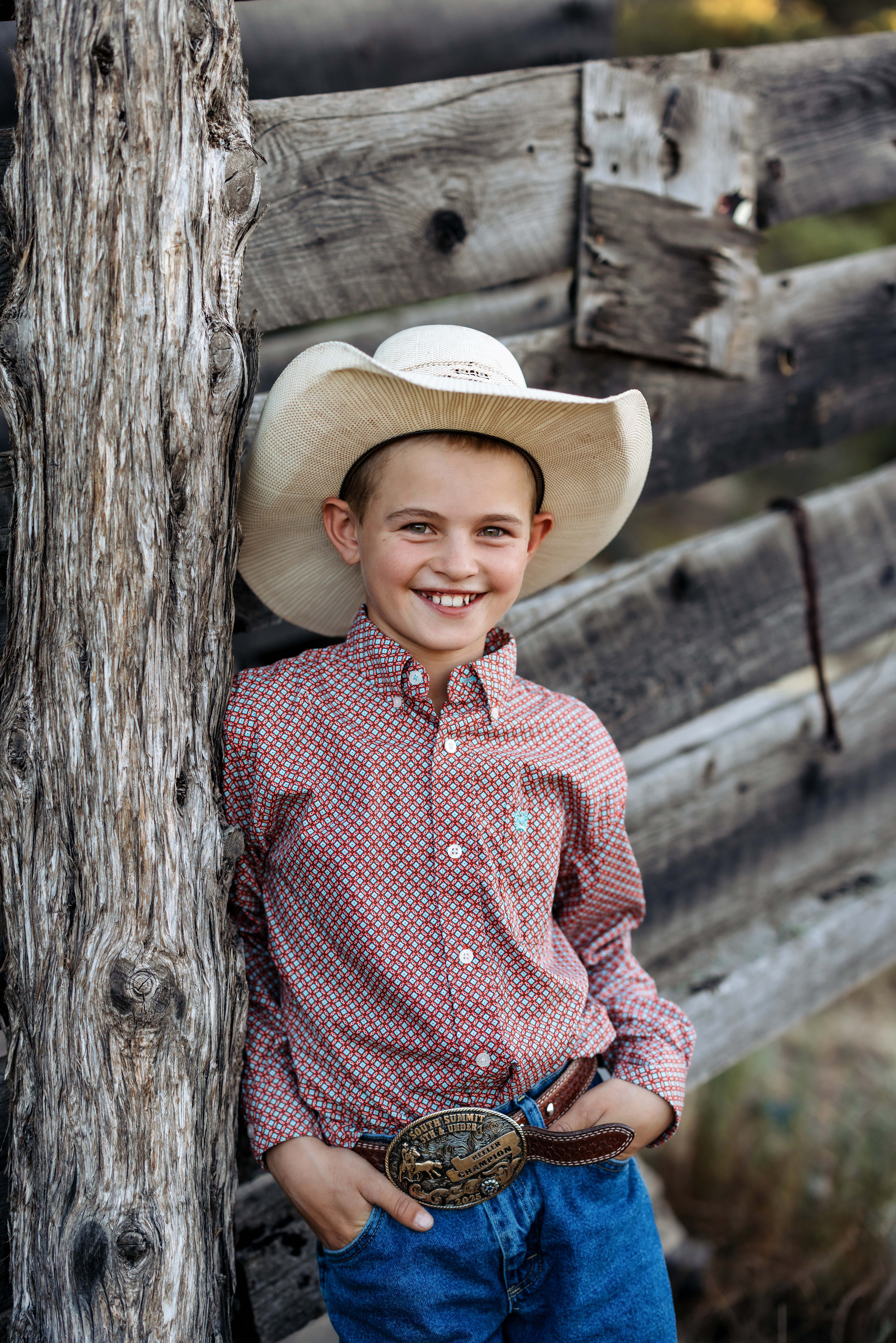 A boy smiles while wearing a cowboy hat, a button up red shirt and a belt buckle